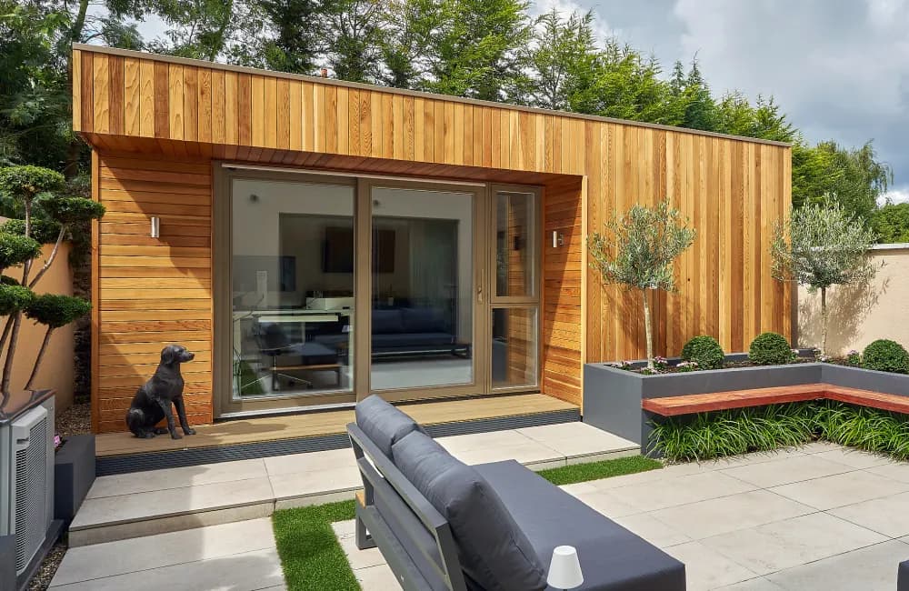 A cedar-clad garden room with sliding glass doors, beside a paved patio with outdoor seating