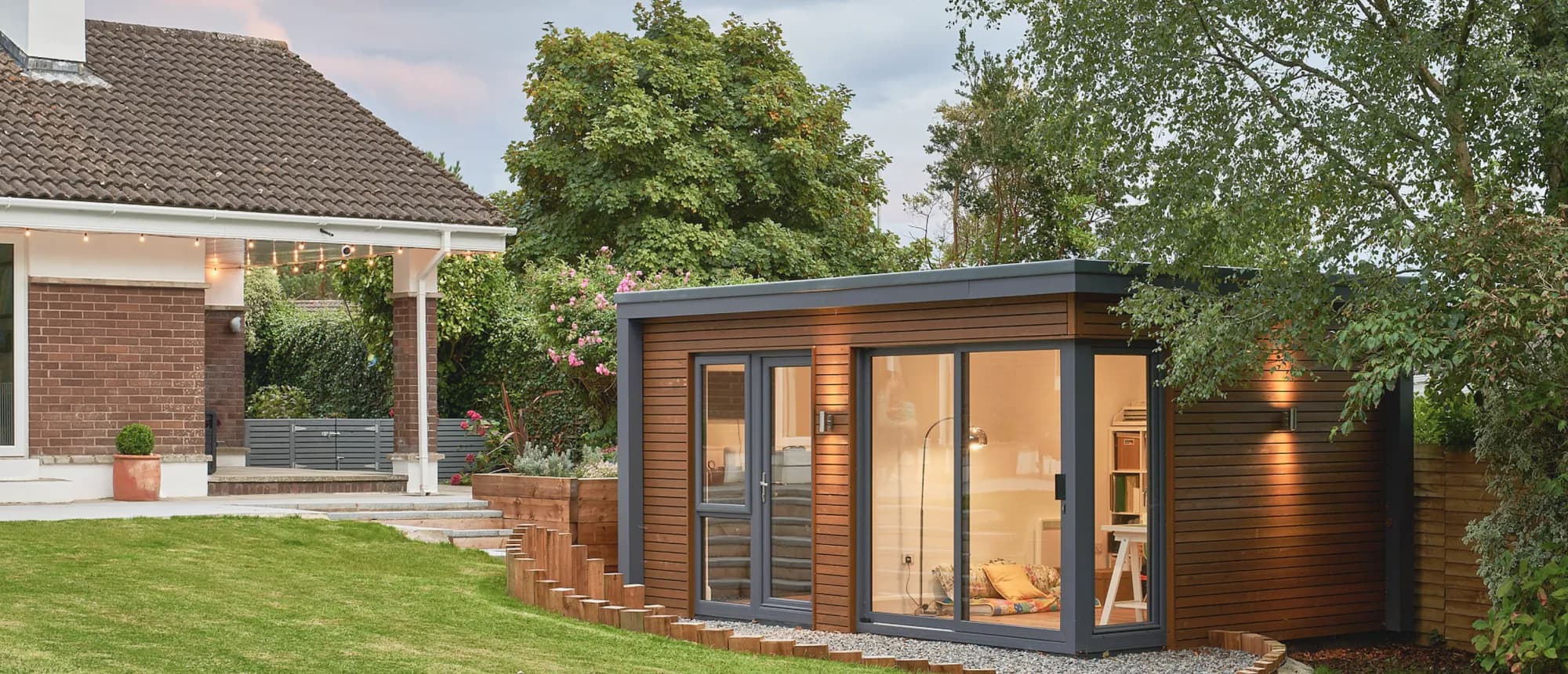 A timber-clad garden room with glass doors in a back garden, beside a house with a covered patio