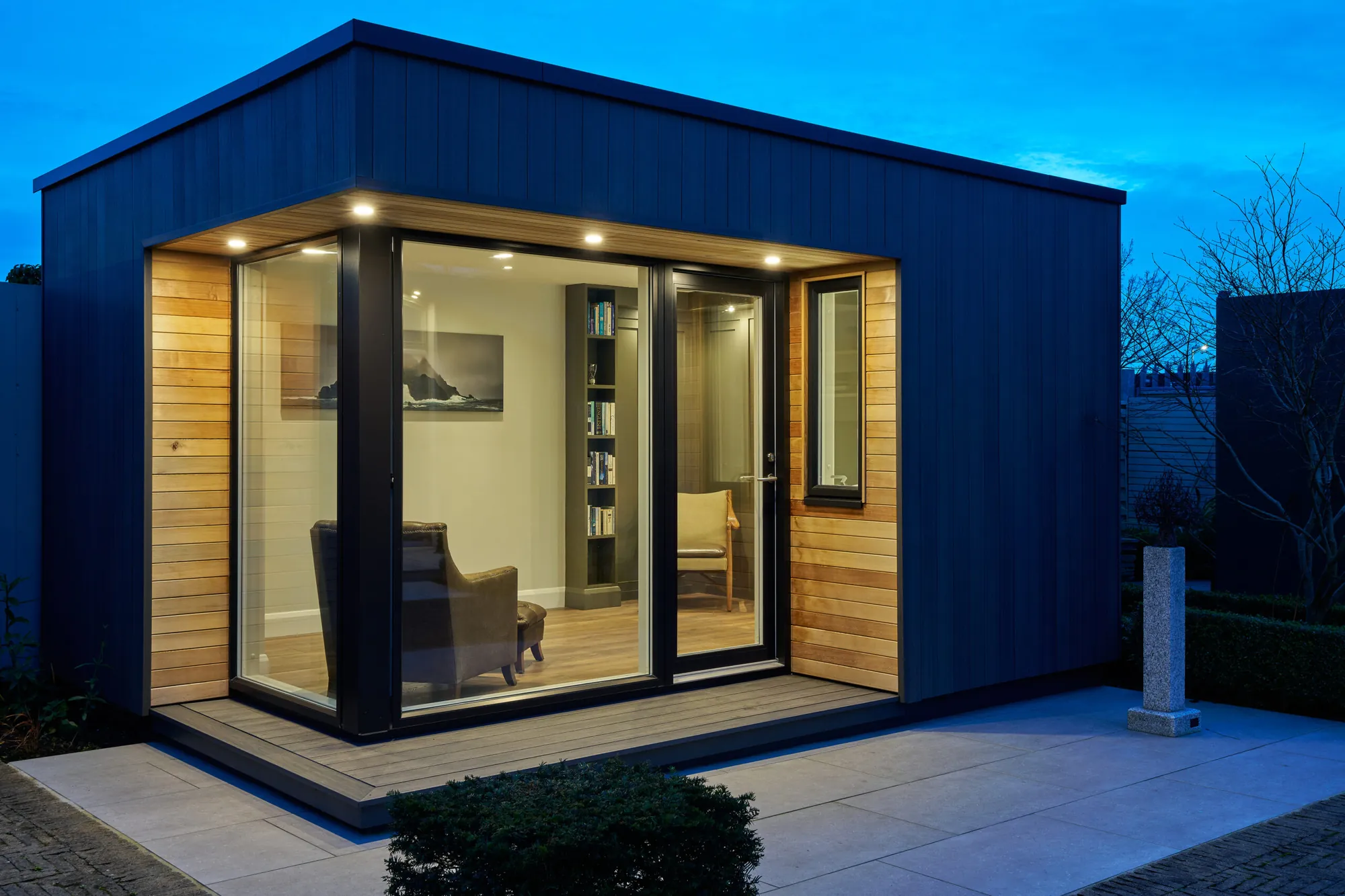 A dark-clad garden room with a cedar-lined glazed corner, lit at dusk