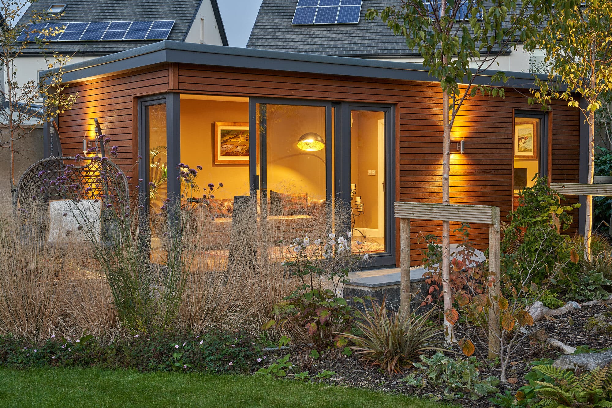 A cedar-clad Shomera Classic Range garden studio lit at dusk, its corner sliding doors glowing over a planted garden