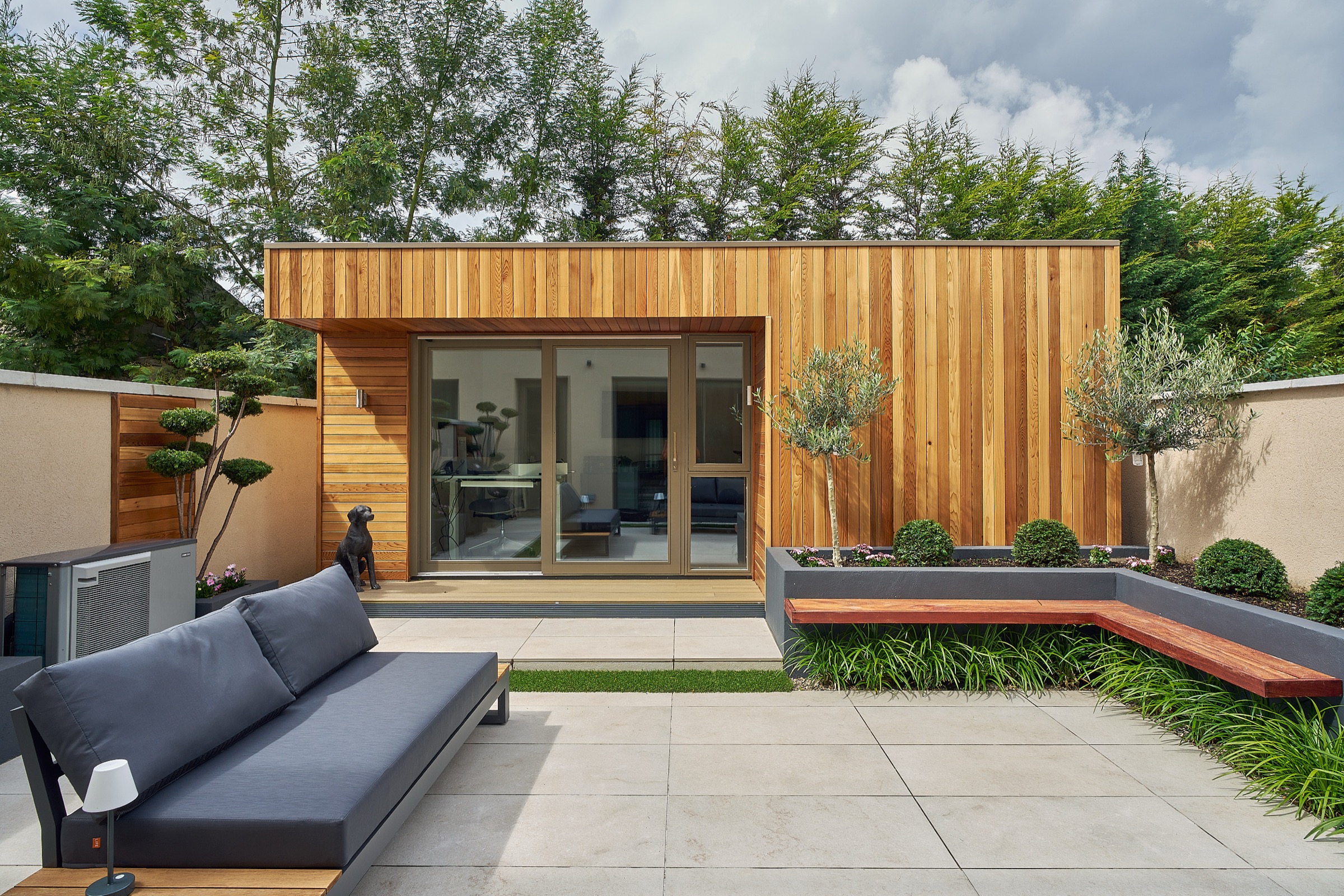 A cedar-clad Shomera garden studio with sliding doors opening onto a paved courtyard with built-in bench seating and planting