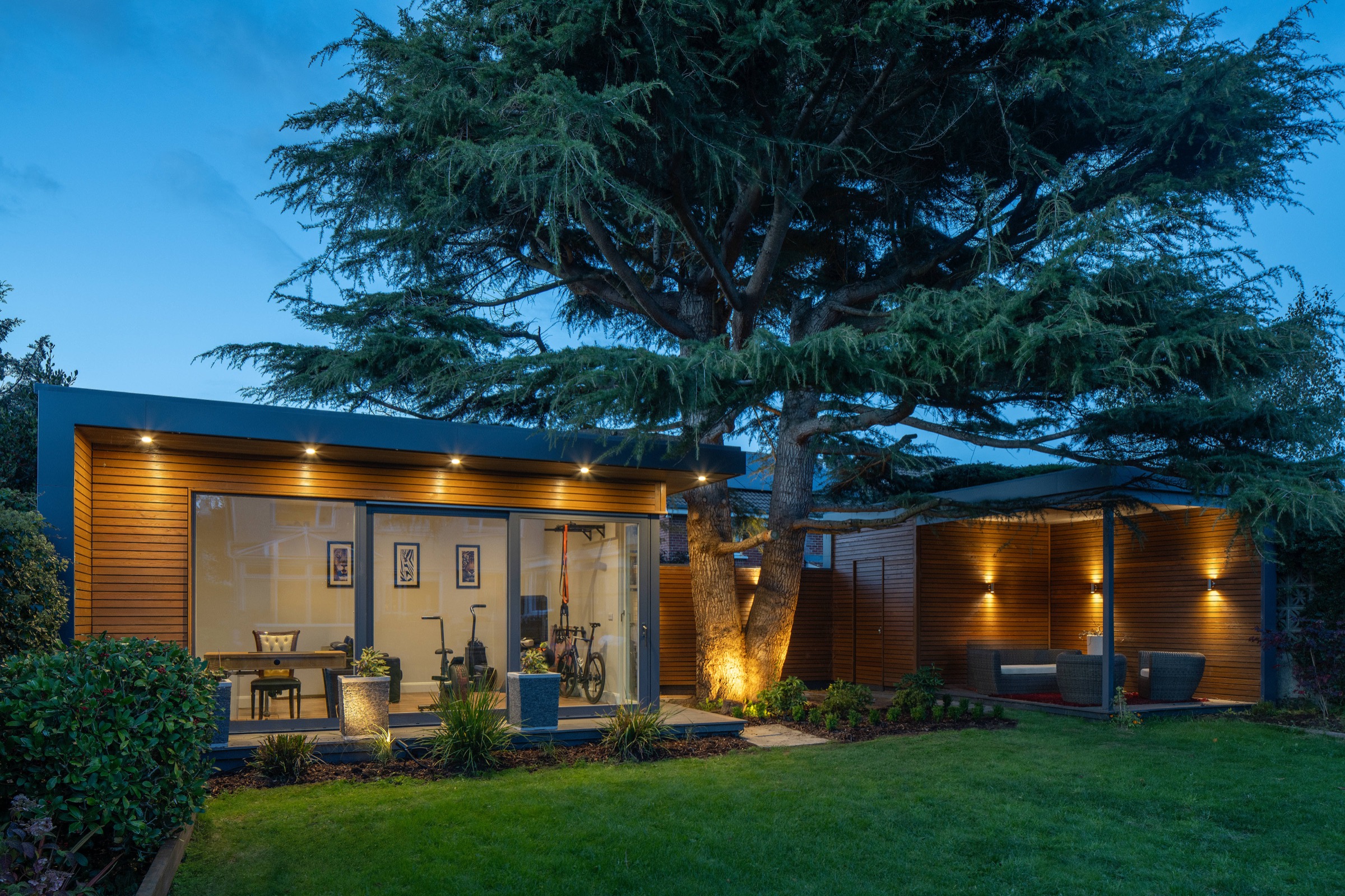A cedar-clad Shomera garden studio used as a home gym, lit at dusk beside a mature cedar tree