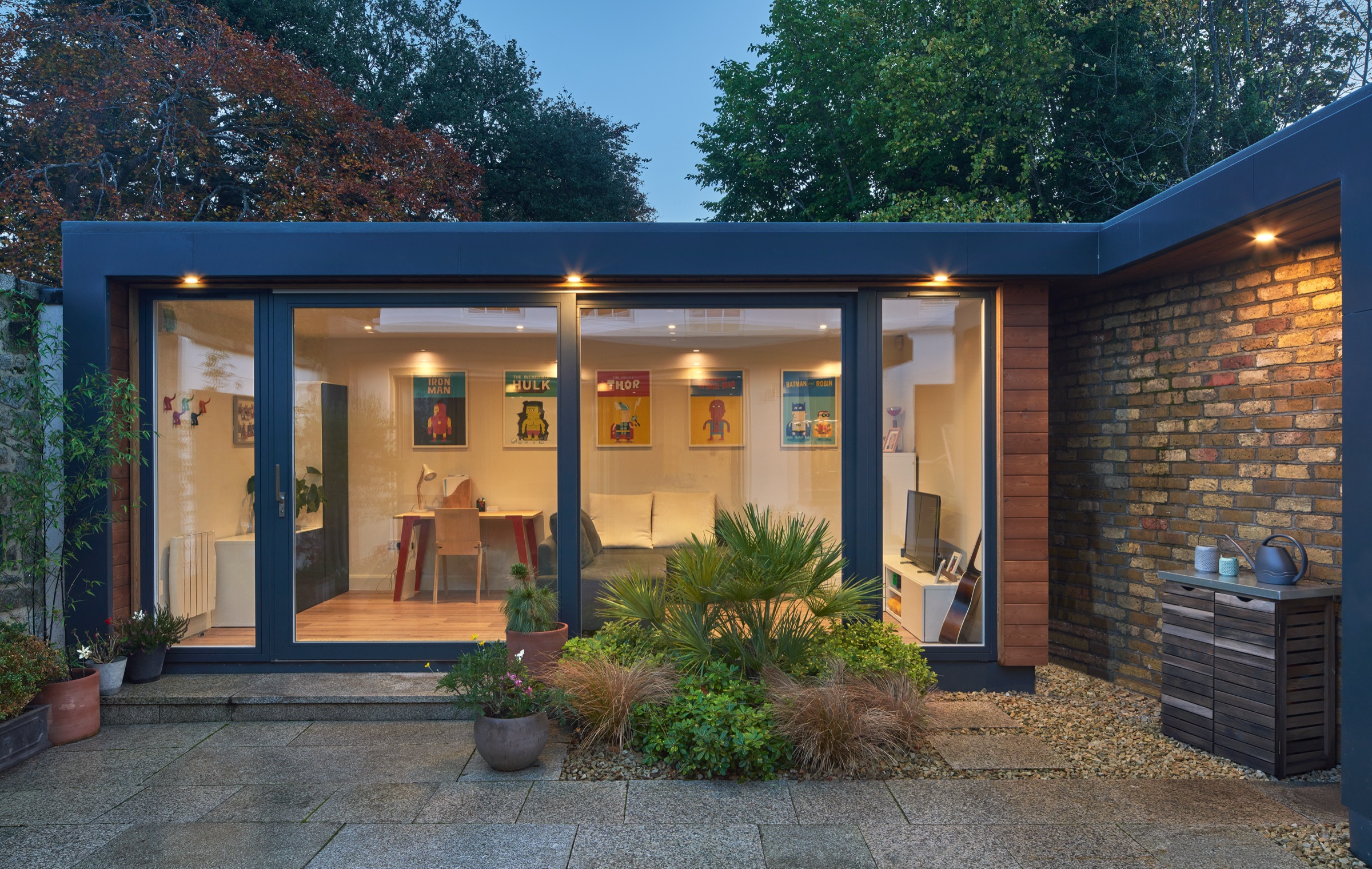 A Shomera garden studio at dusk, set up as a home office with a desk and a sofa beyond