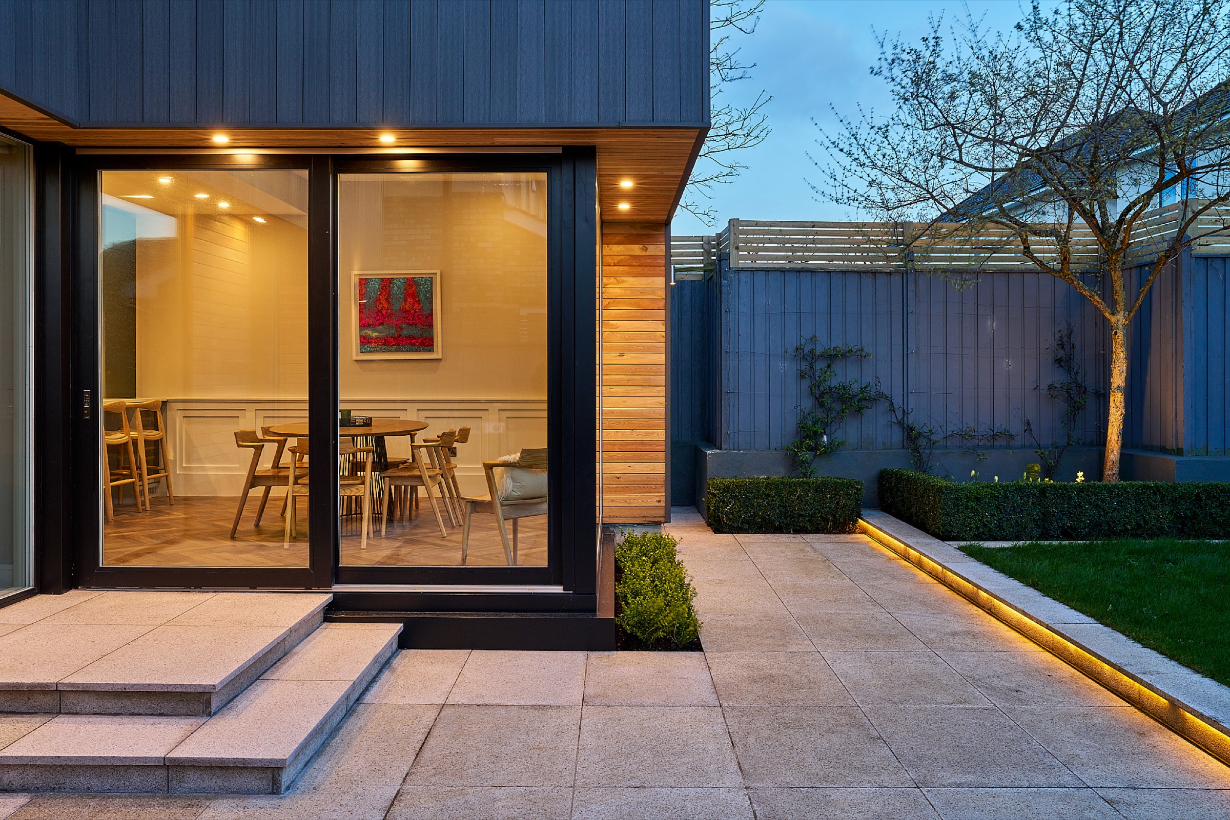 A Shomera house extension at dusk, glazed sliding doors opening onto a warmly lit dining room