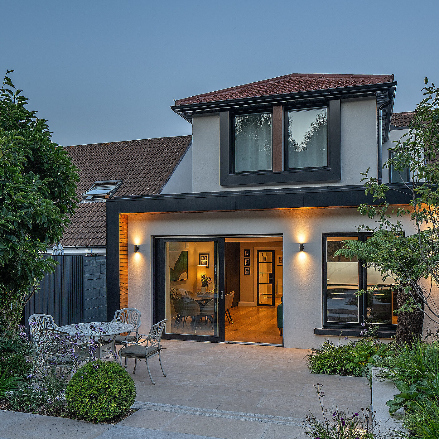 A Shomera house extension lit at dusk, its glazed doors open onto a paved patio beneath a first-floor extension