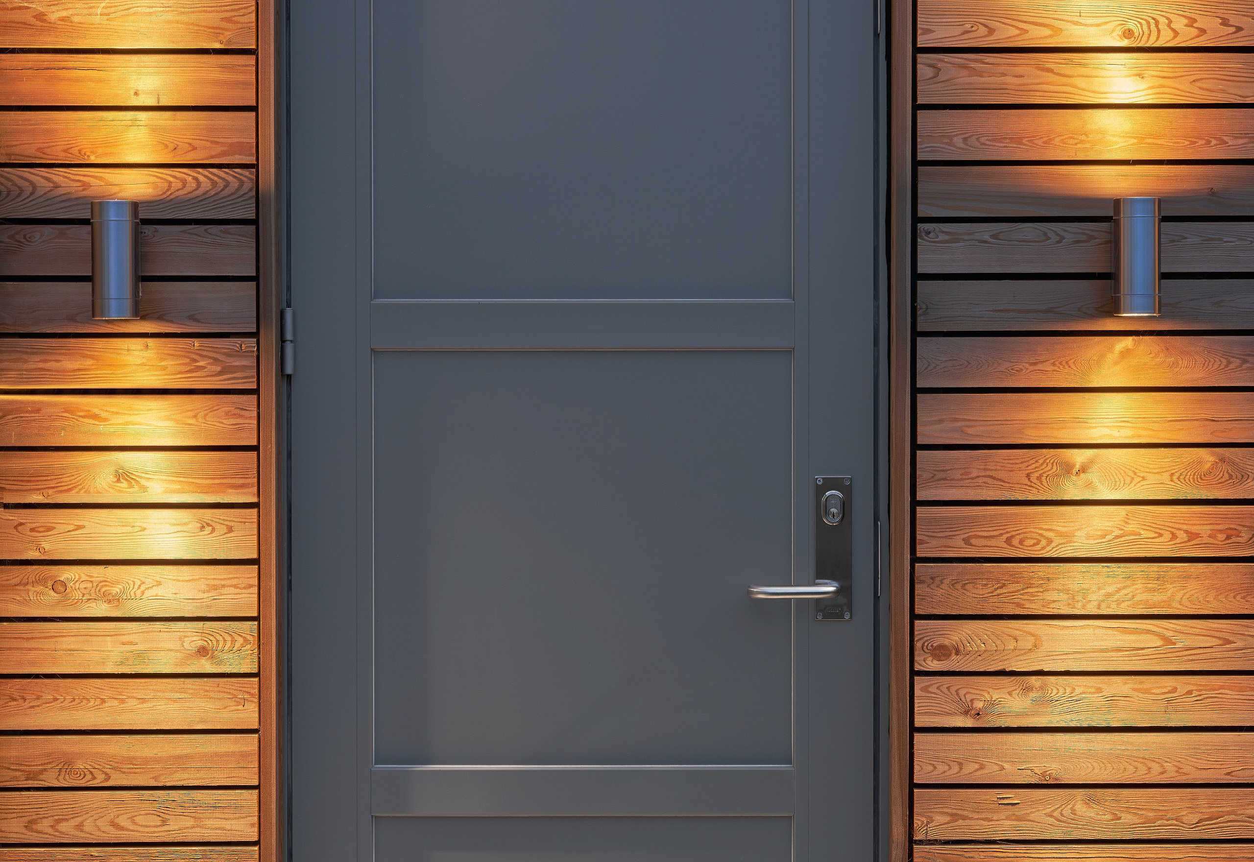 The studio's grey entrance door, flanked by uplit cedar cladding