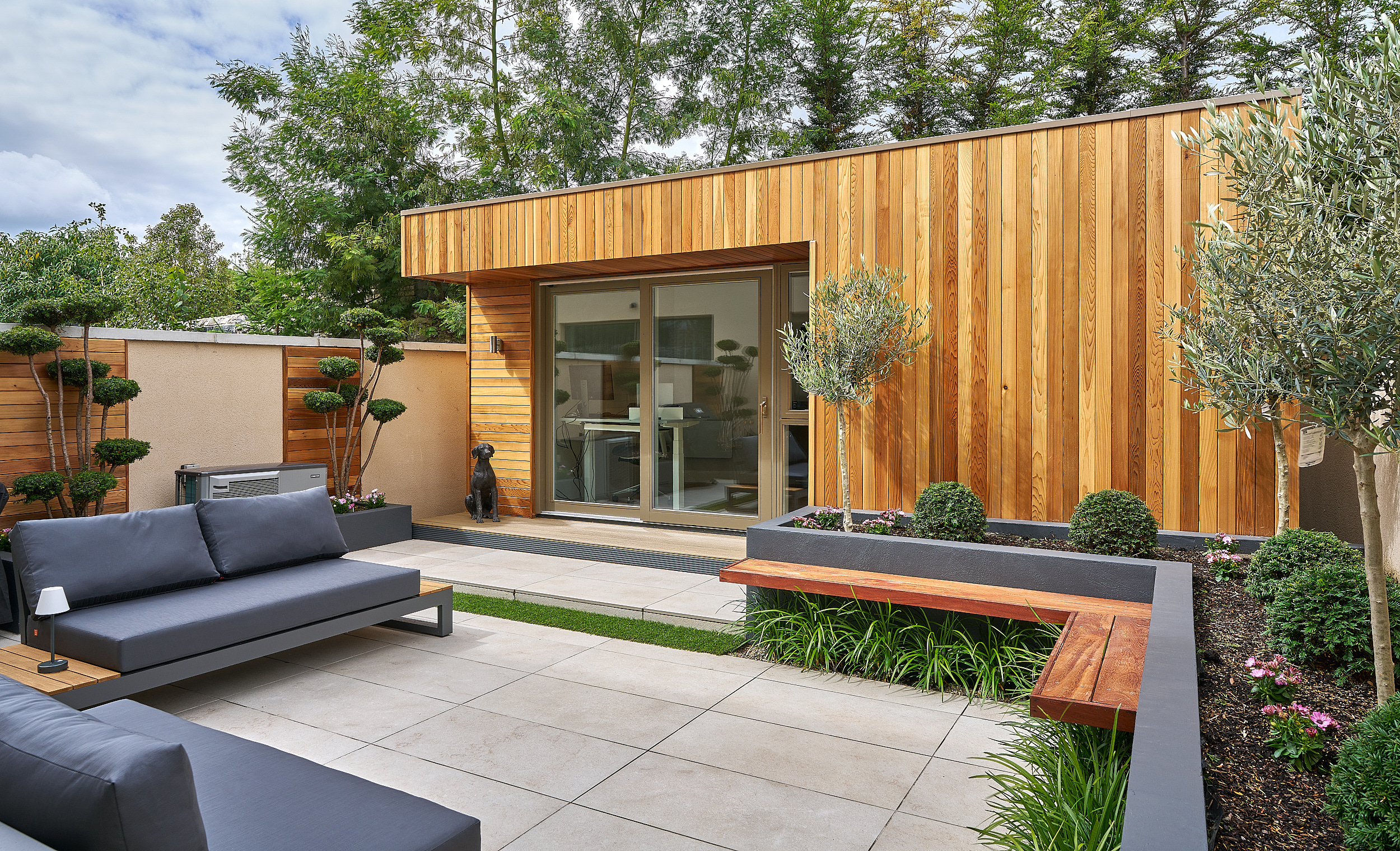 The cedar-clad garden studio at the end of the paved terrace, with an olive tree and built-in seating