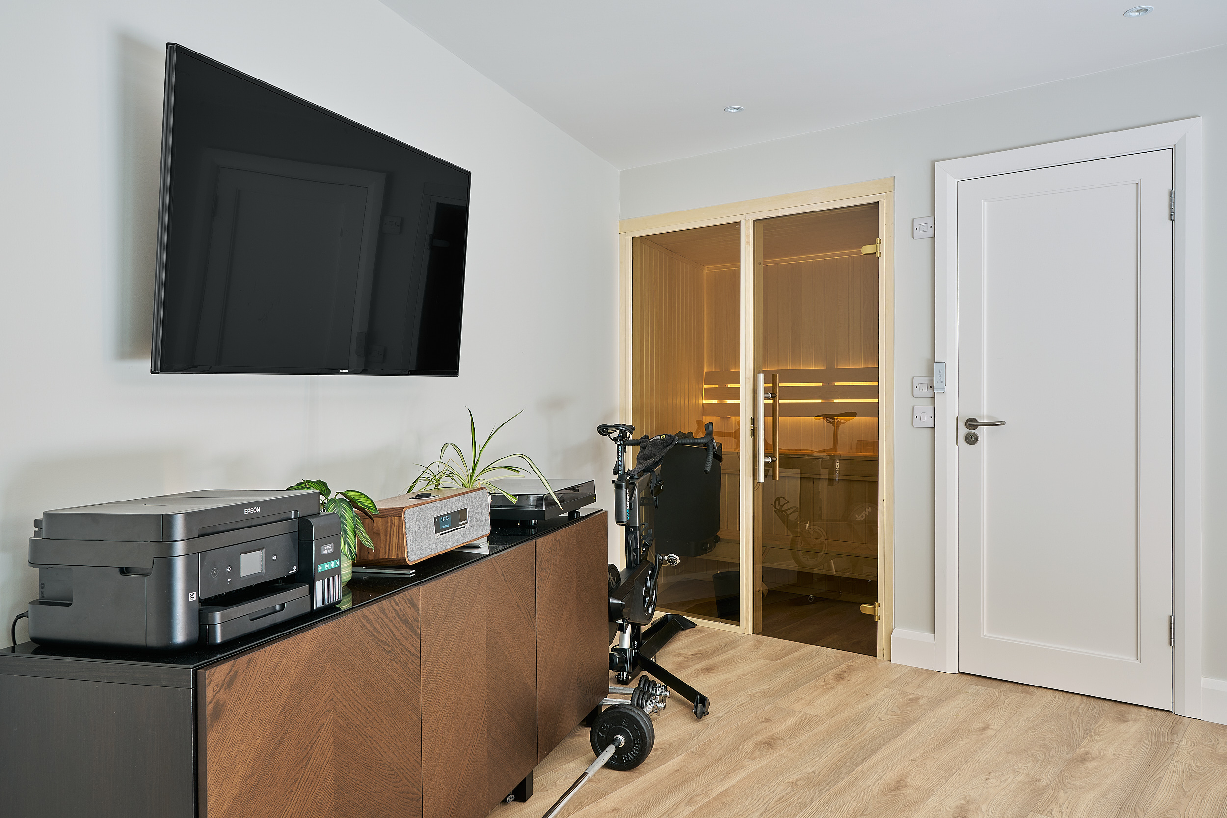 Inside the studio, the glazed sauna door beside a wall-mounted screen and gym equipment