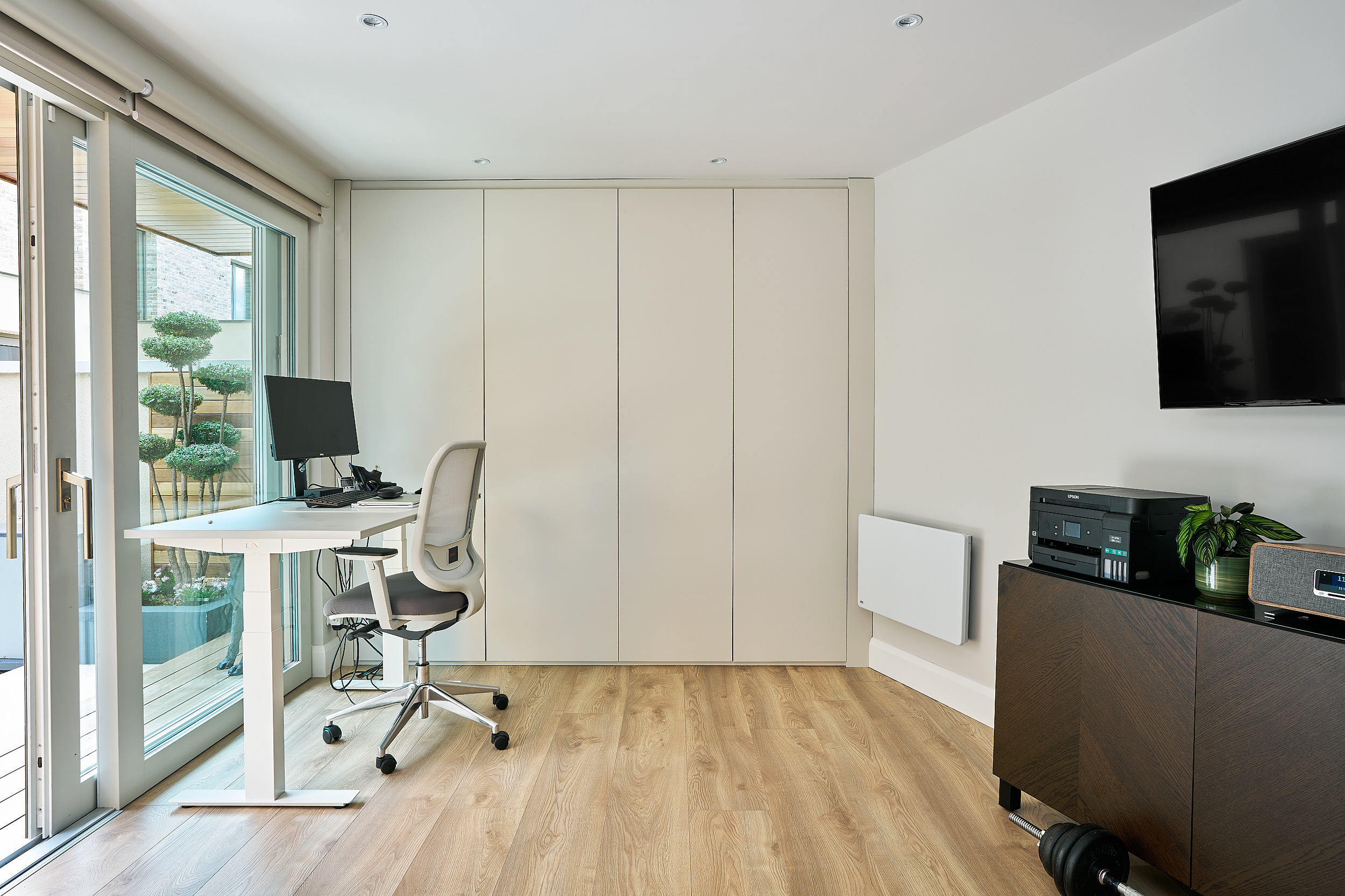The studio's office zone, a height-adjustable desk beside full-height glazing and fitted storage