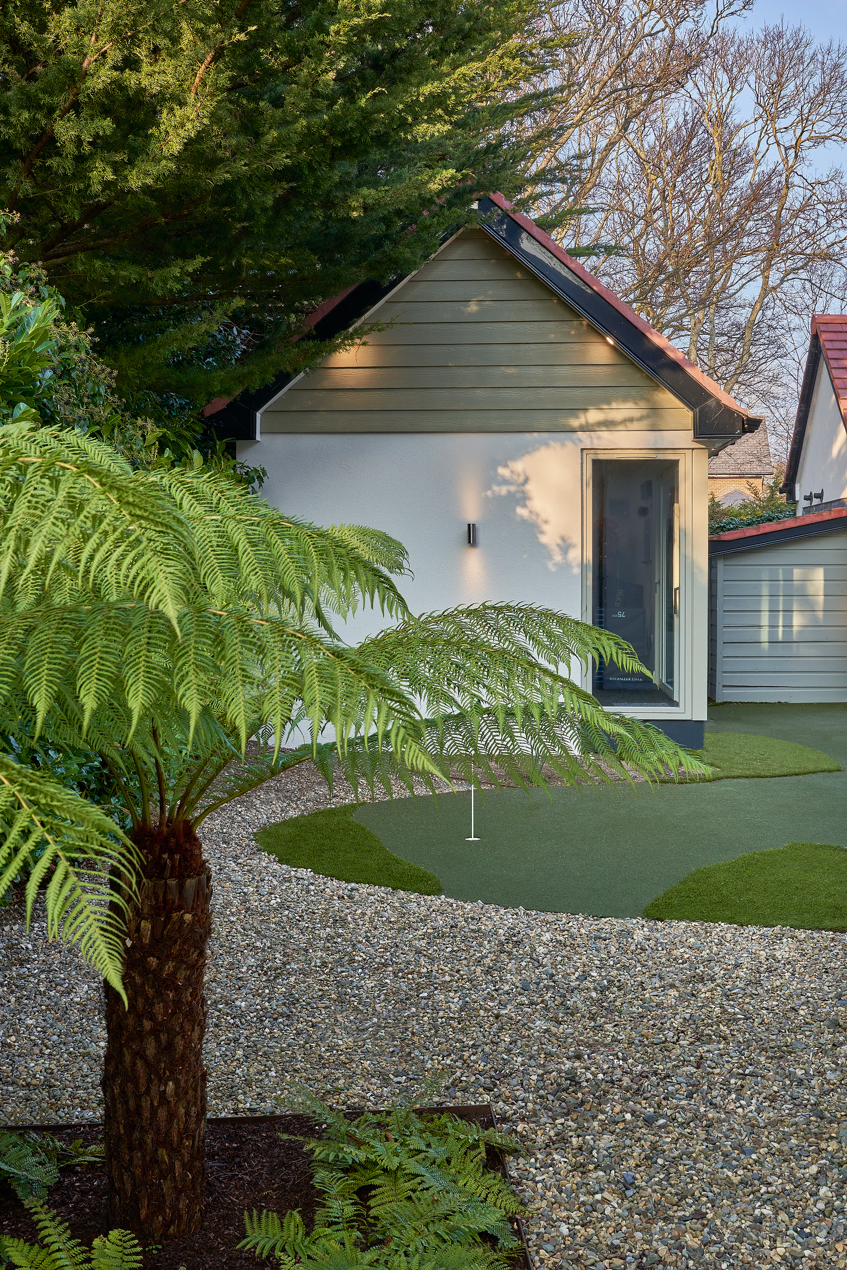A mature tree fern in the foreground, the studio beyond across gravel and lawn