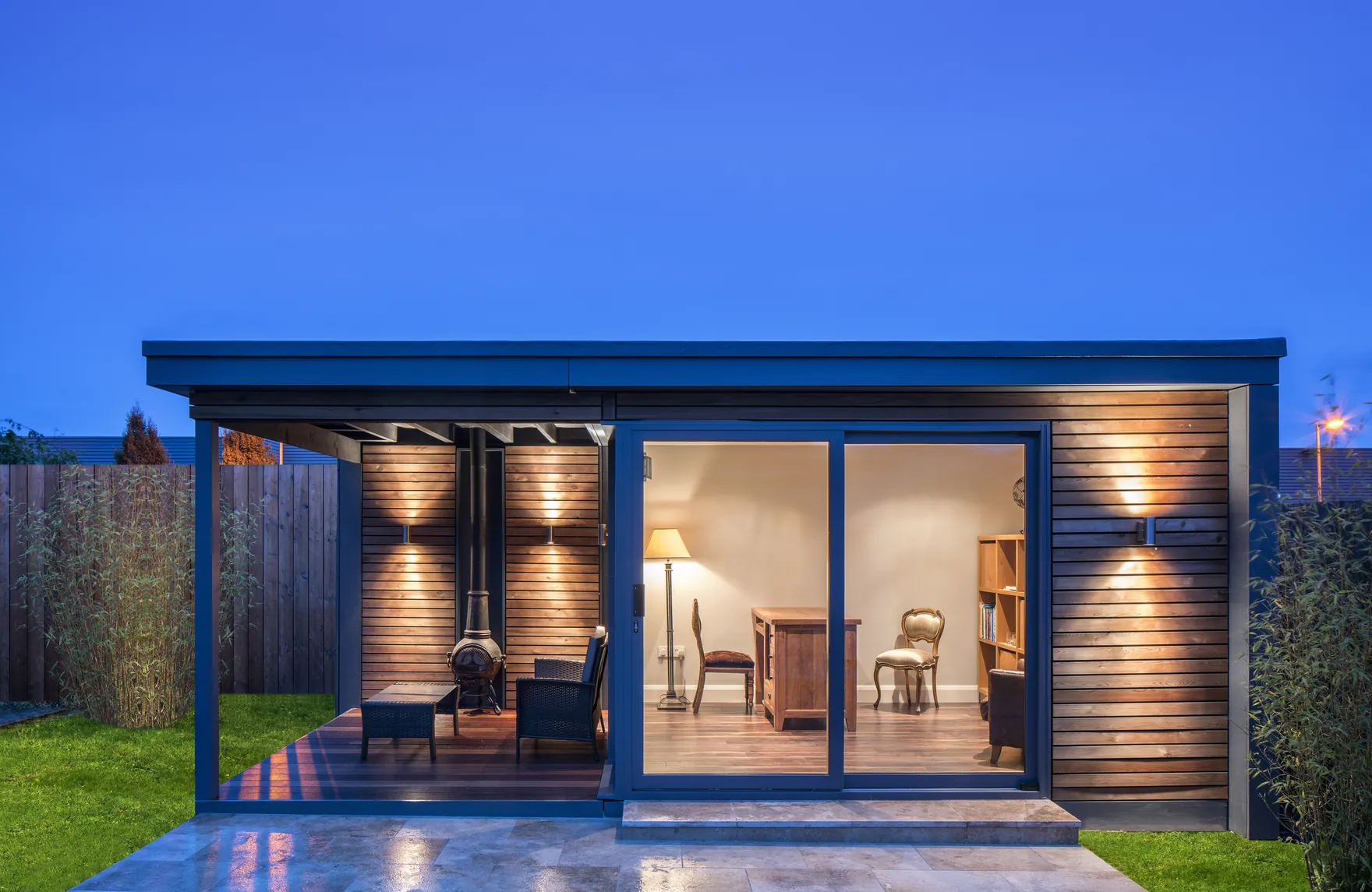 A garden room with a covered deck and an outdoor stove, its sliding doors open onto a home office, at dusk