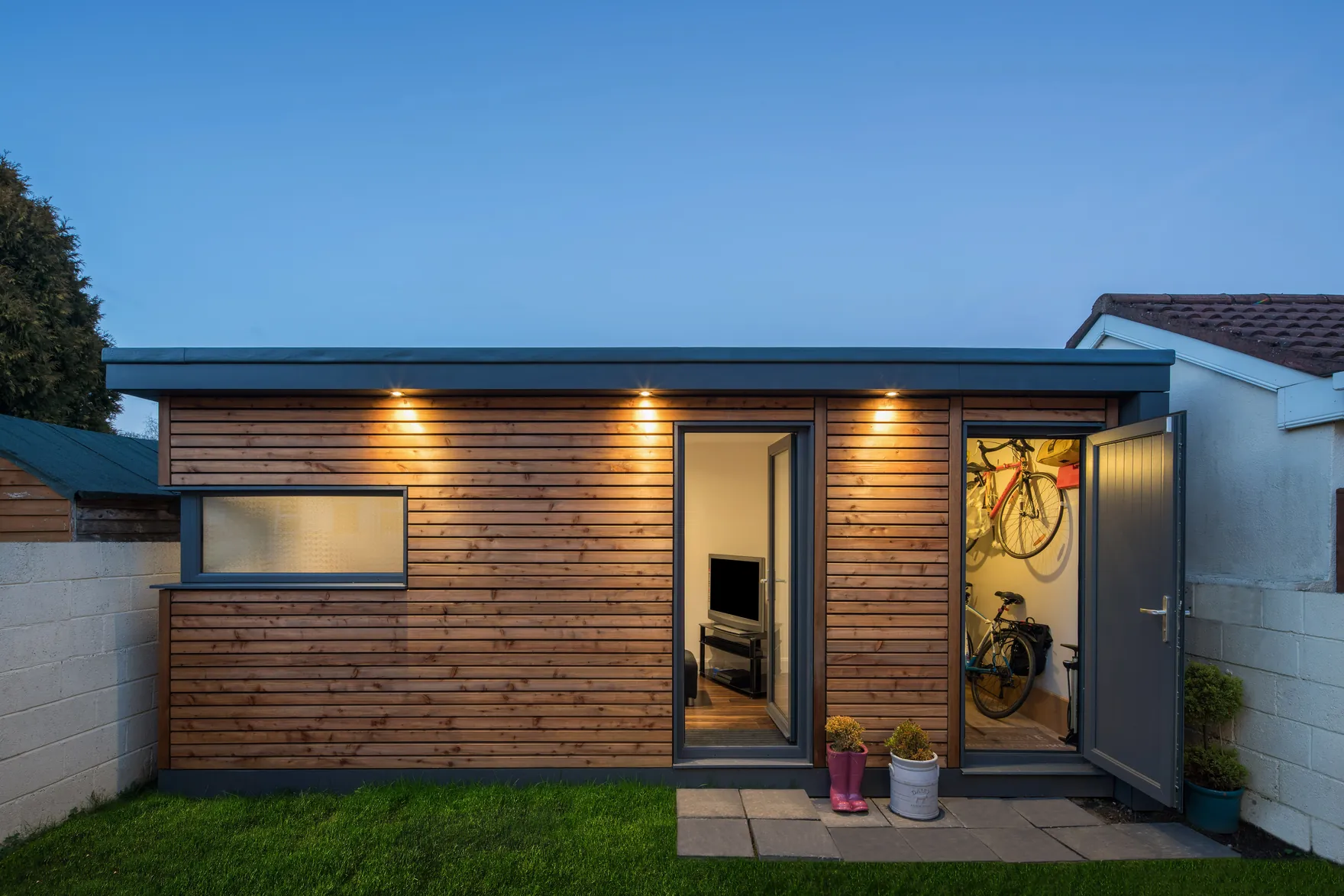 A timber-clad garden room with its doors open, showing bicycles stored inside, at dusk