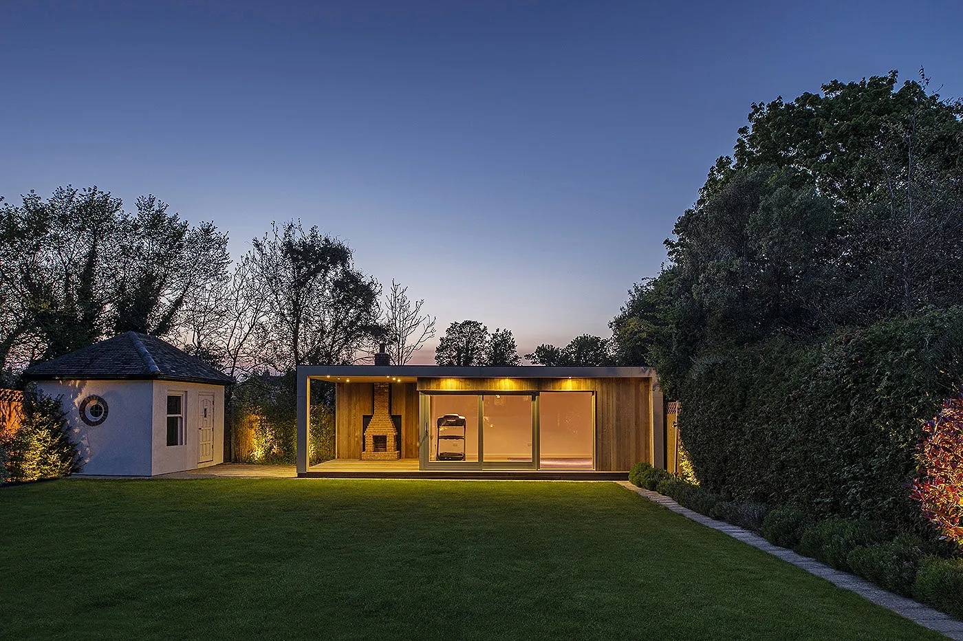 A garden room with a covered deck beside a white garden building, at the end of a long lawn at dusk