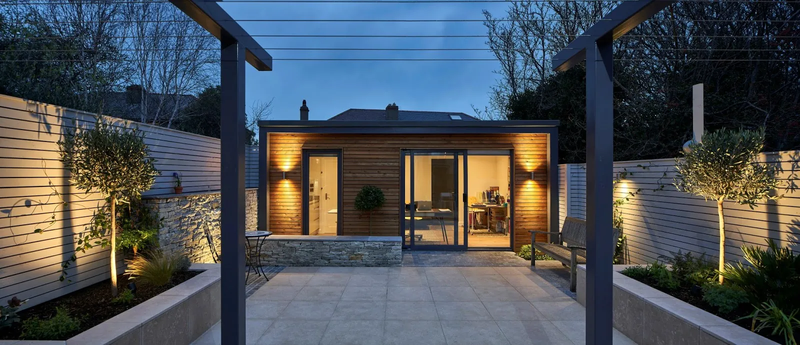 A timber-clad garden room lit at dusk, seen across a paved courtyard between two pergola posts