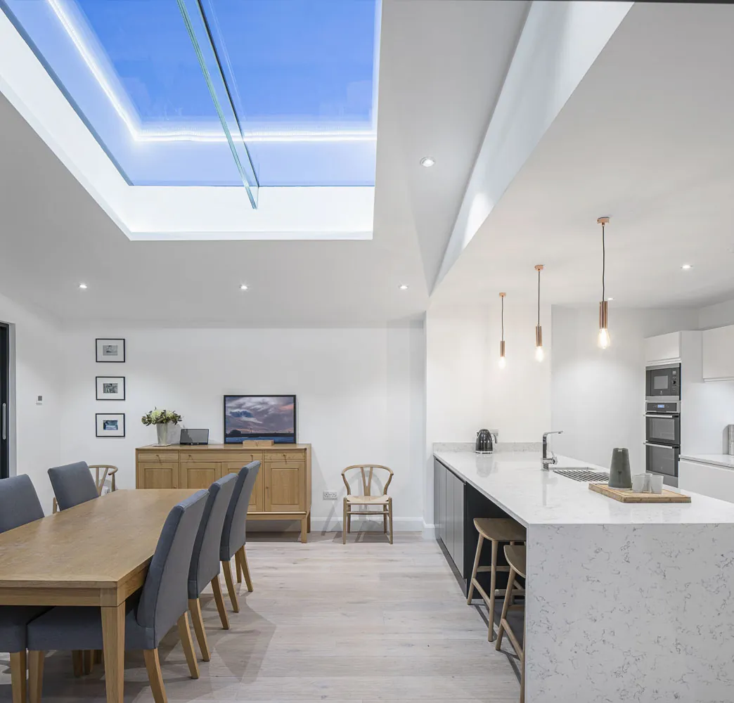 A kitchen island with a white stone worktop beside a dining table, under a large rooflight