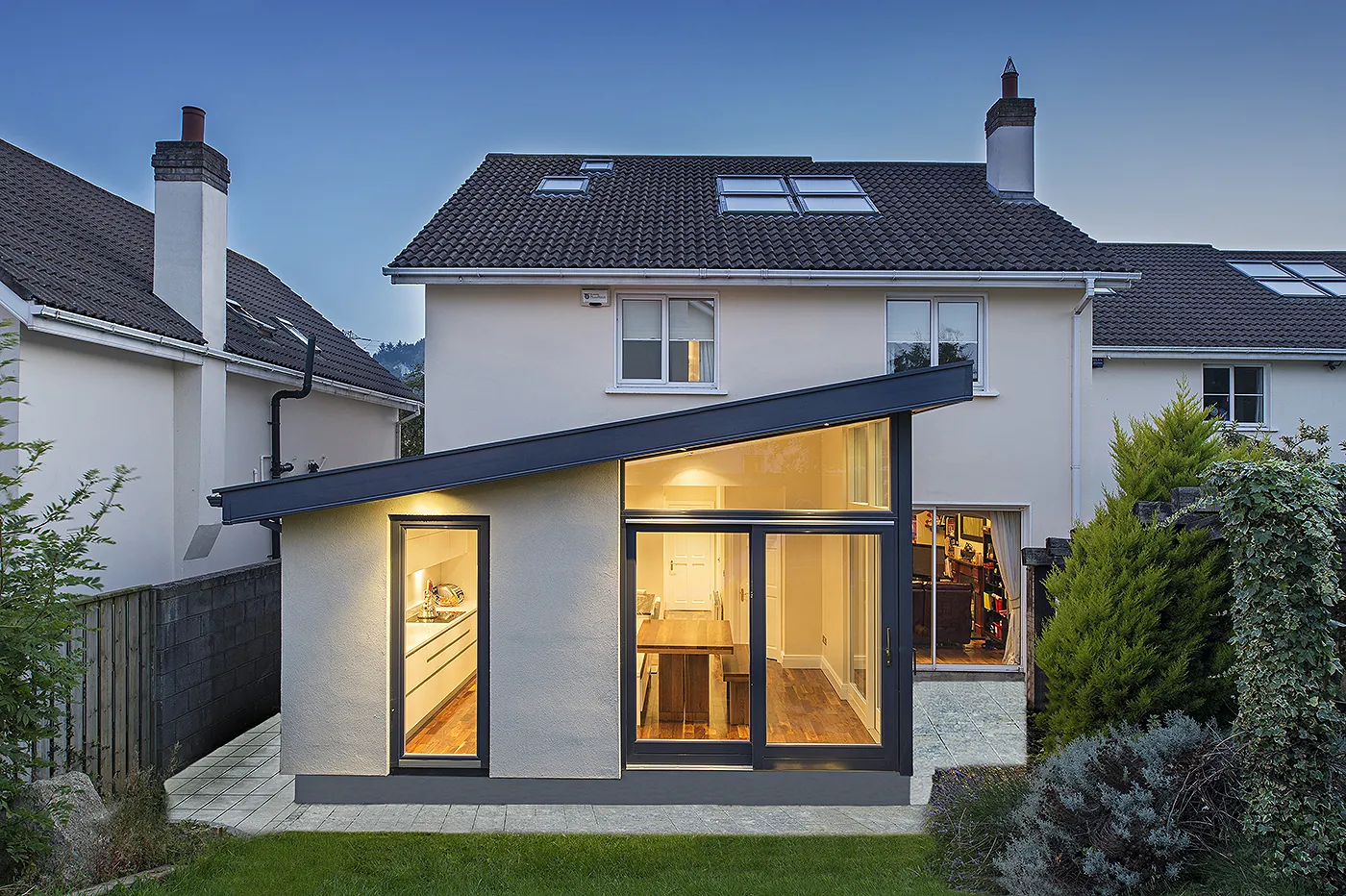 A rendered house extension with a mono-pitch roof and tall glazing, at the back of a two-storey house