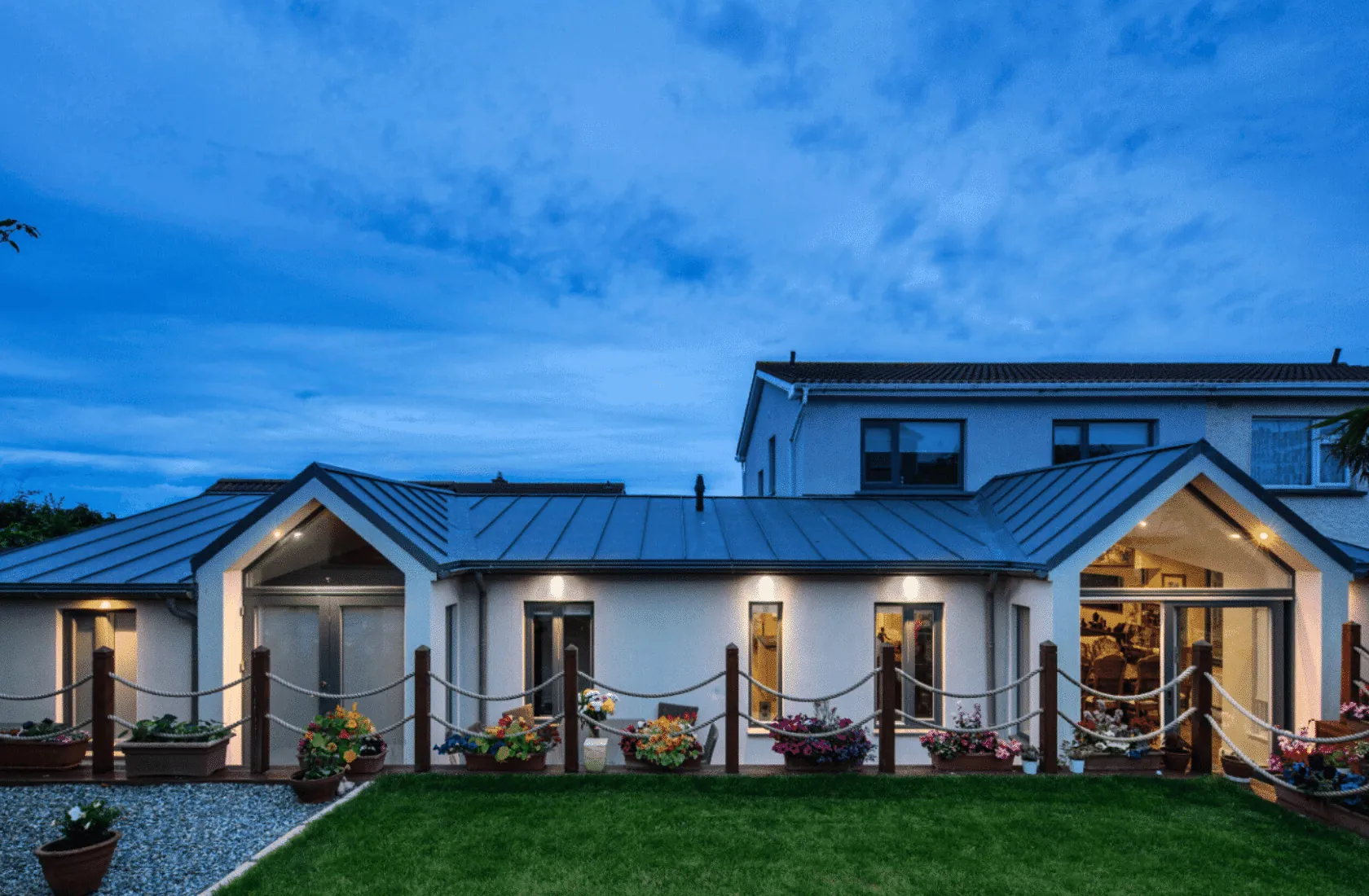 A single-storey extension with a zinc roof and two glazed gables across the back of a house, at dusk