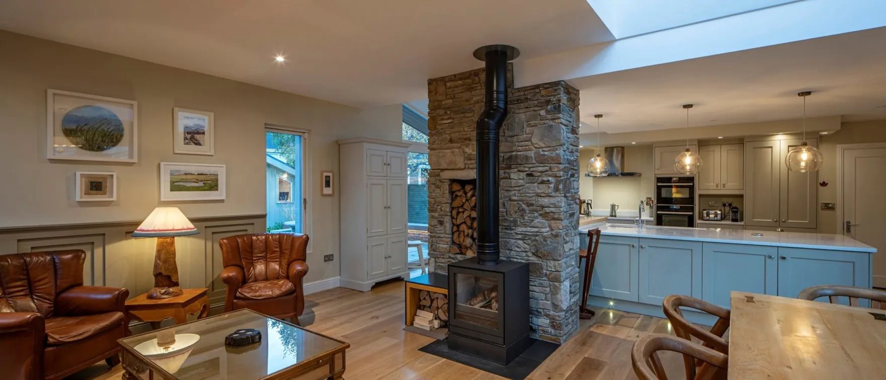 An open-plan living area and kitchen divided by a stone chimney with a wood-burning stove