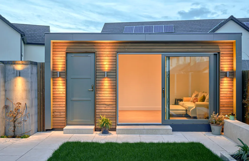 A timber-clad garden room with a grey door and sliding glass doors, lit at dusk