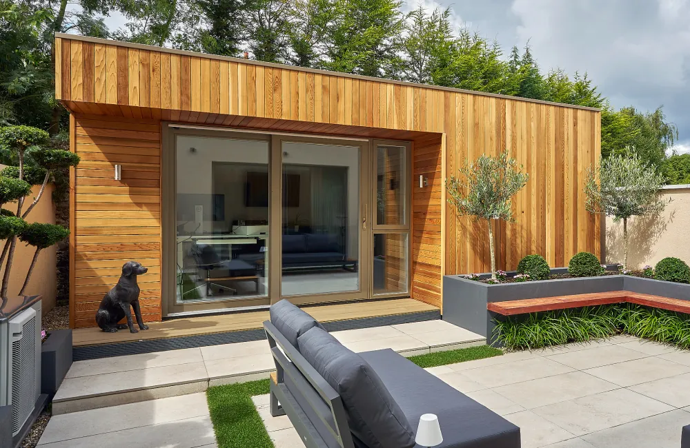 A cedar-clad garden room with sliding glass doors, beside a paved patio with outdoor seating