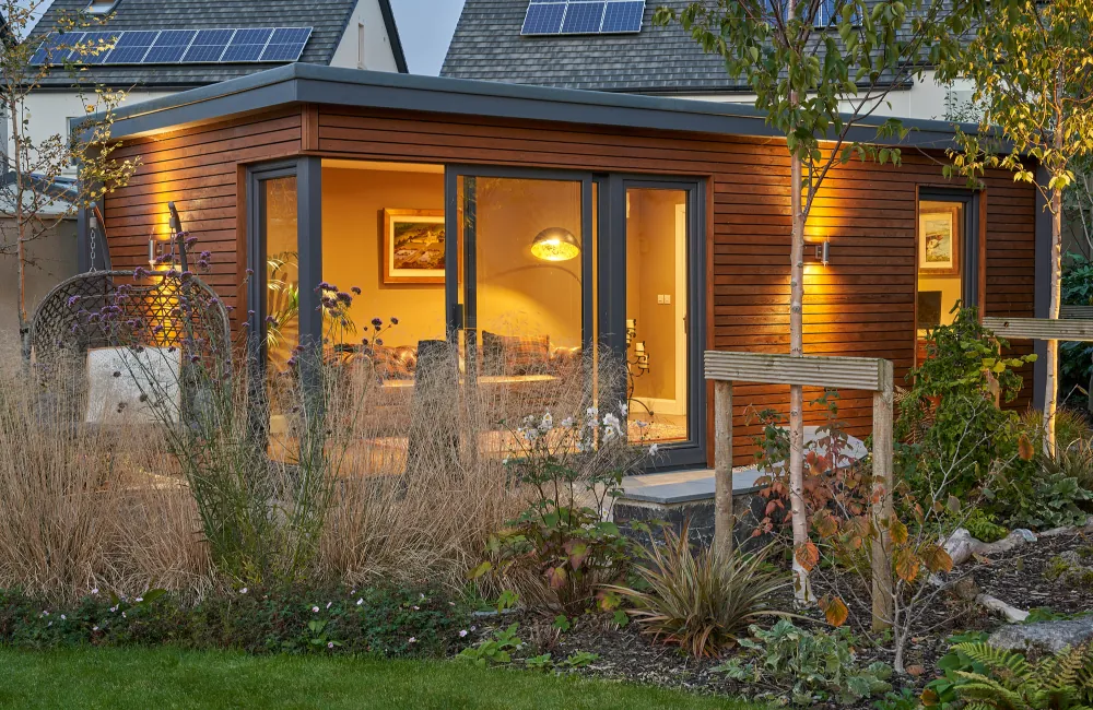 A timber-clad garden room with corner glazing, lit from inside, behind garden planting
