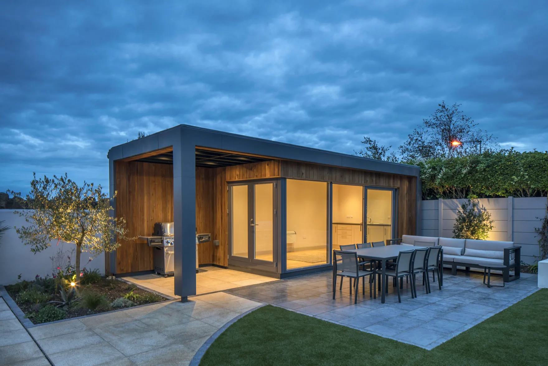 A timber-clad garden room with a covered canopy at dusk, with a barbecue and an outdoor dining table on the patio