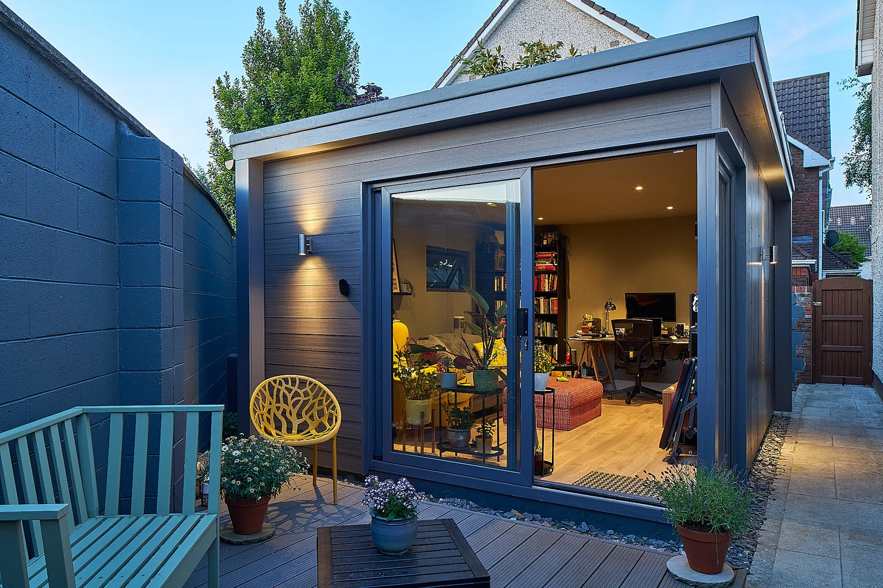 A grey garden room with its sliding door open onto a home office, beside a small deck with a bench