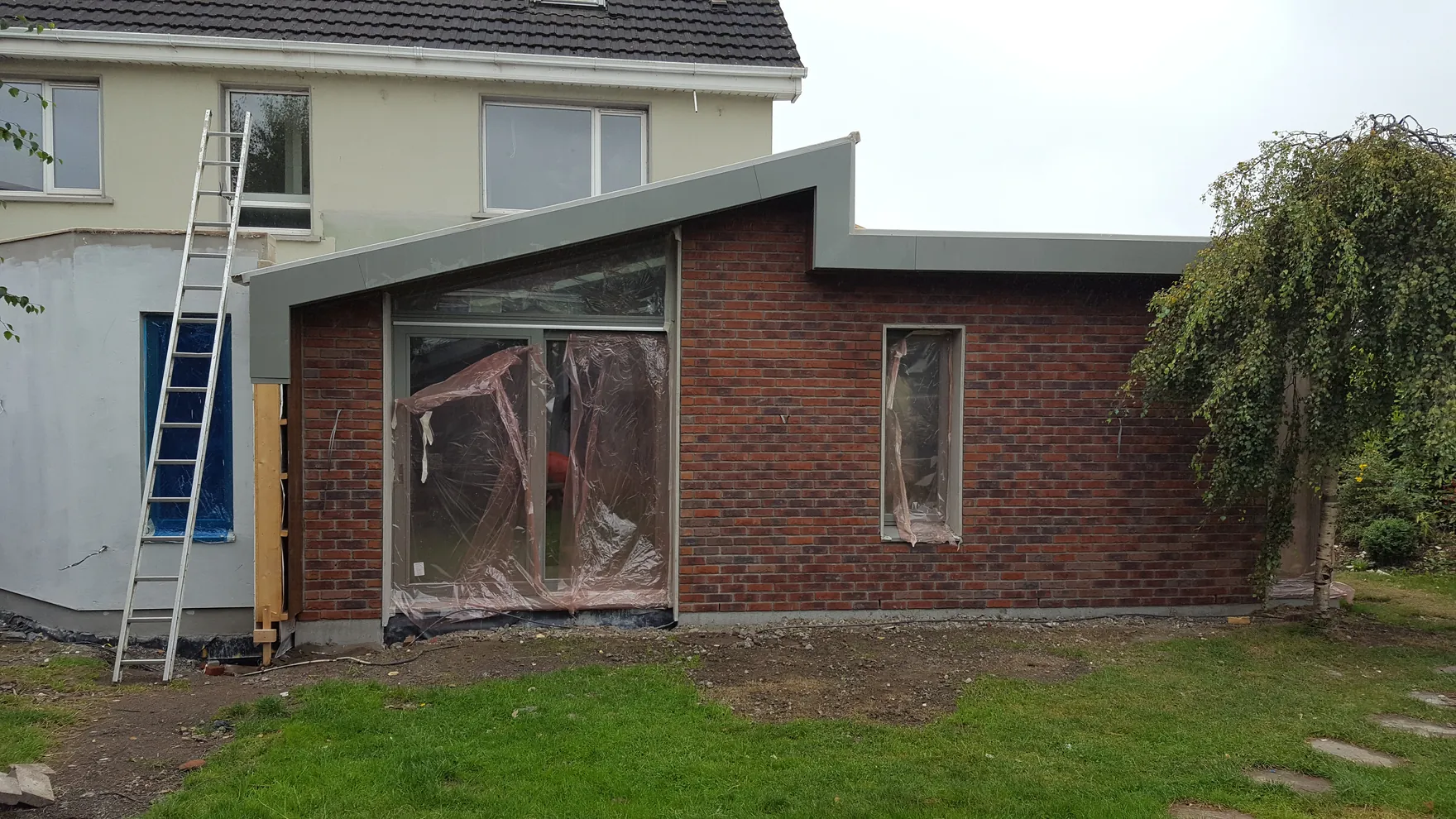A brick house extension under construction, with a ladder against the house and plastic sheeting over the window openings