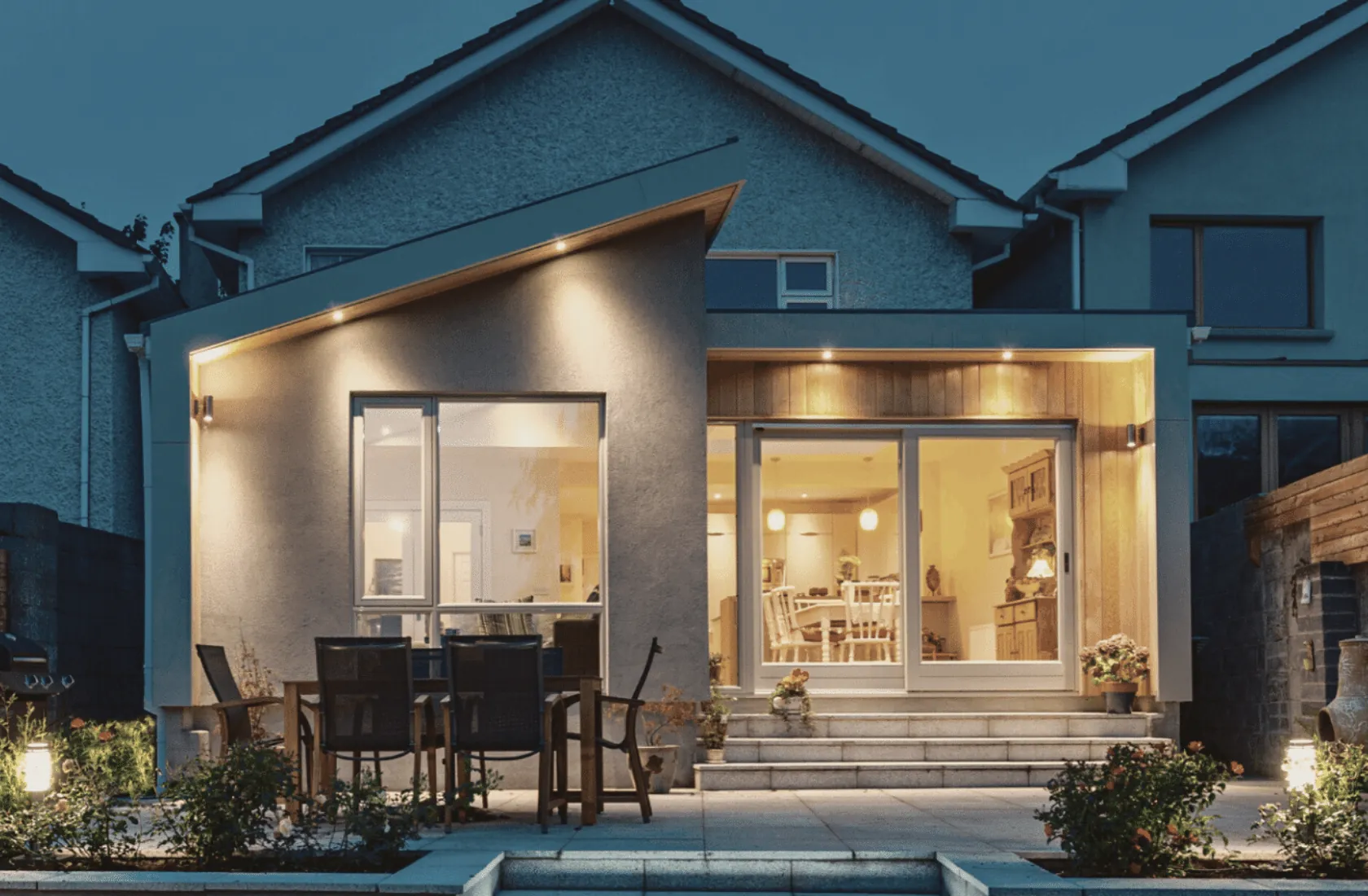 A house extension with a sloping roof and a timber-clad recess, lit at dusk above a terraced patio