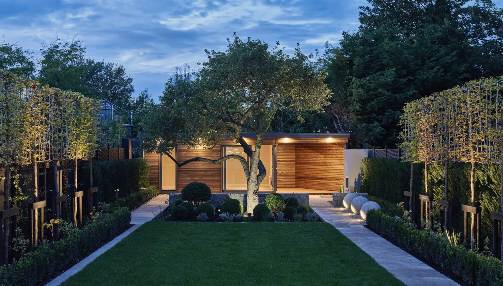 A garden room at the end of a long lawn, behind a mature tree, lit at dusk