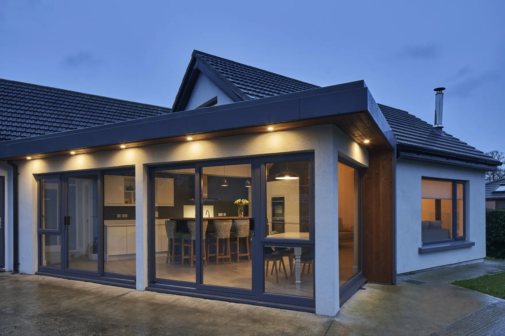 A single-storey house extension with floor-to-ceiling glazing and a kitchen inside, at dusk