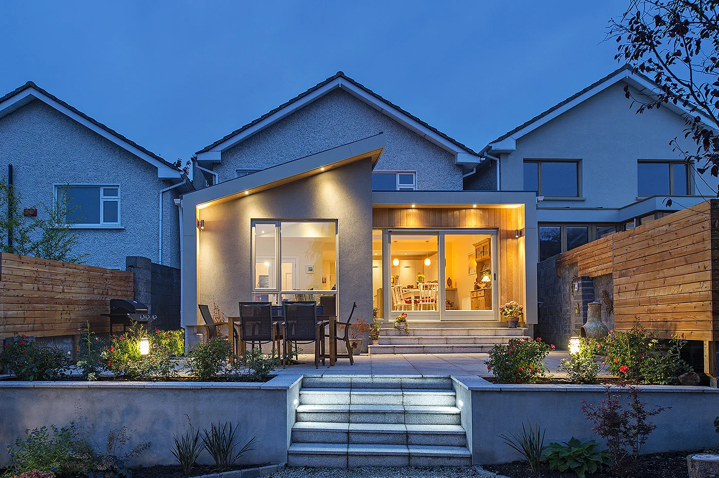 A house extension with a sloping roof and a timber-clad recess, lit at dusk above a terraced patio