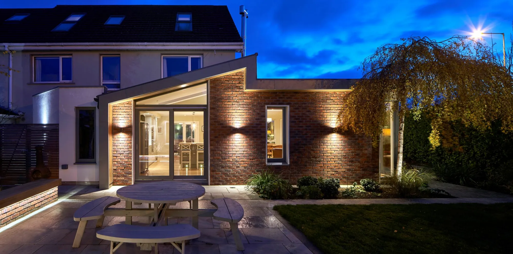 A brick house extension with an angled roof, lit at dusk, with a round picnic table on the patio