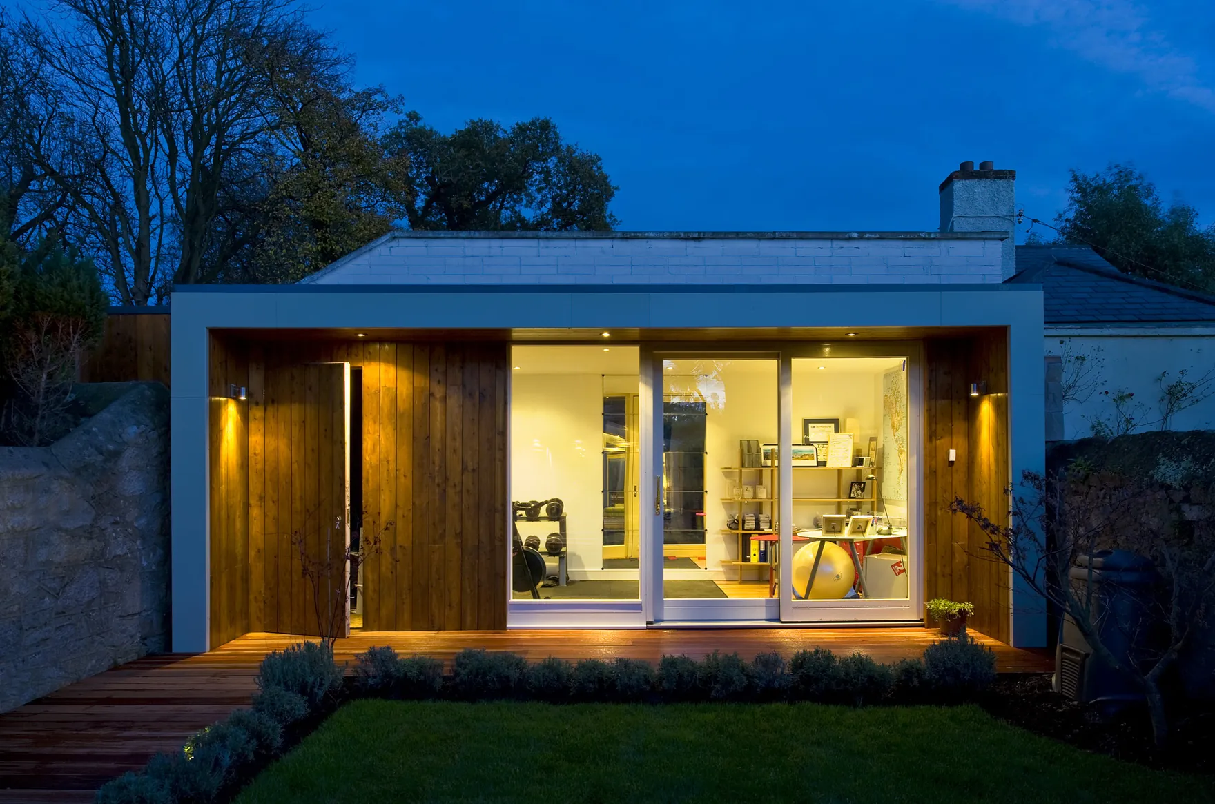 A garden room with timber cladding and glass doors, used as a home gym, lit at dusk