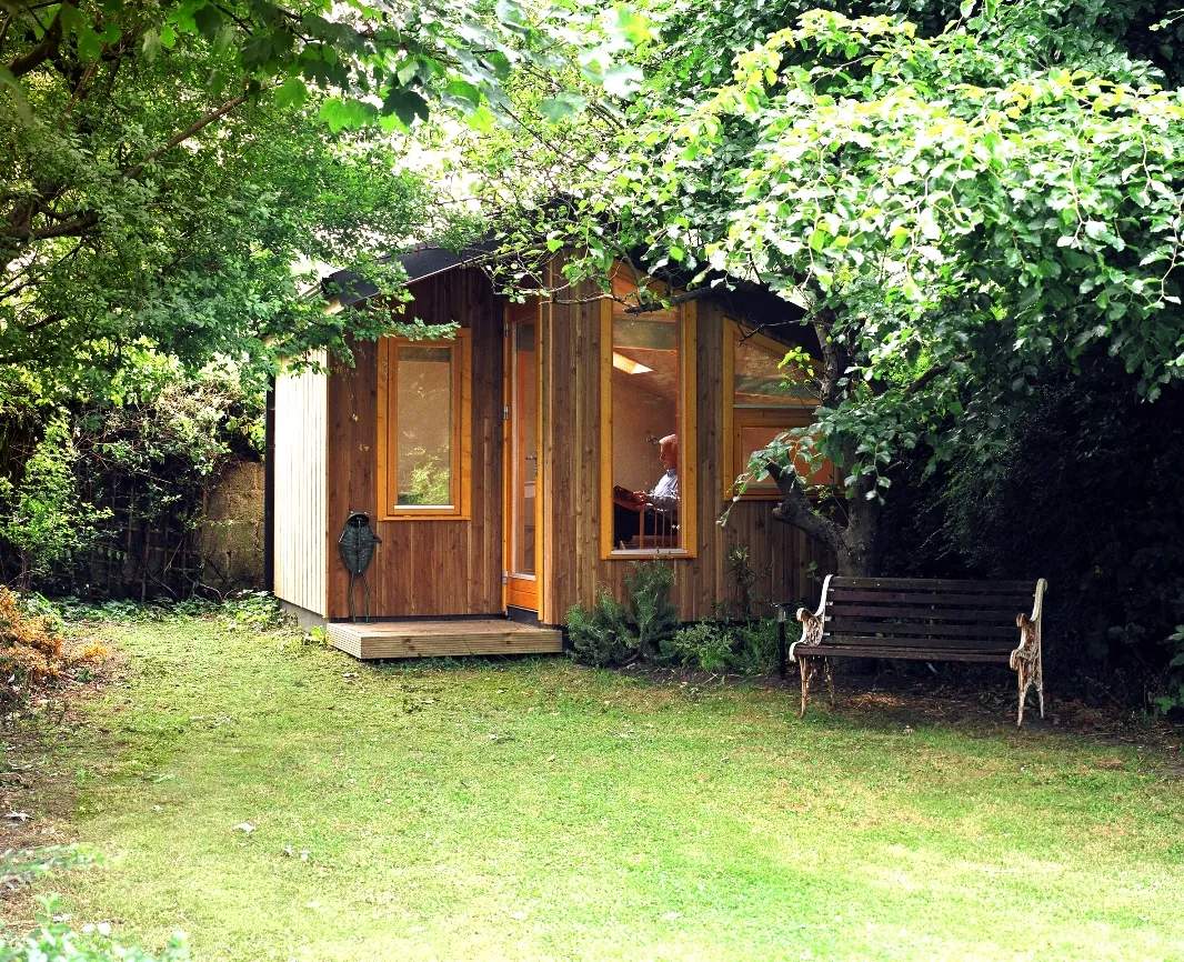 A small timber garden room under trees at the end of a lawn, with a bench beside it