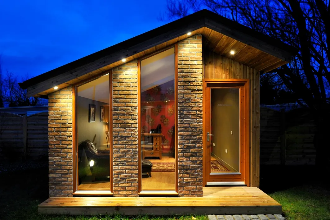A small garden room with brick piers, tall windows and a pitched roof, lit at dusk