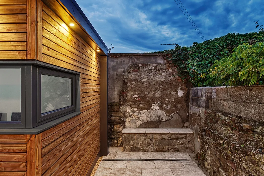 The corner of a timber-clad garden room beside an old stone wall and a small paved courtyard