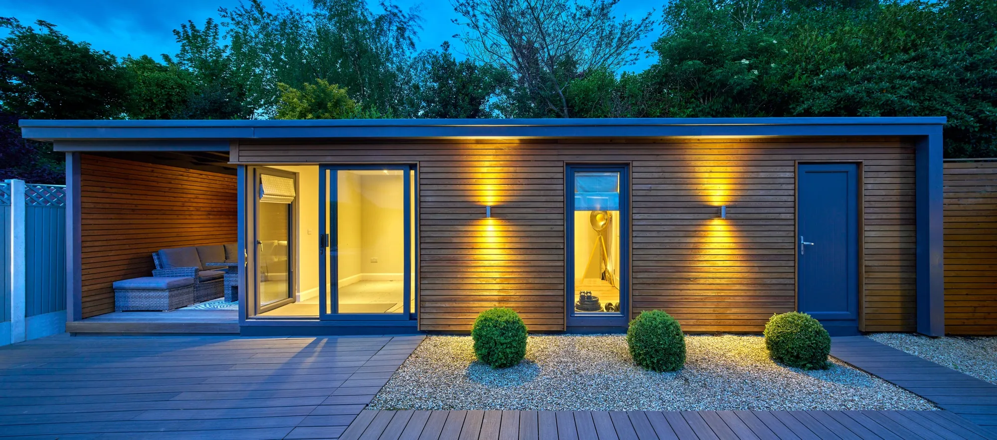 A long timber-clad garden building with a covered seating area, lit at dusk beside a deck and a gravel bed