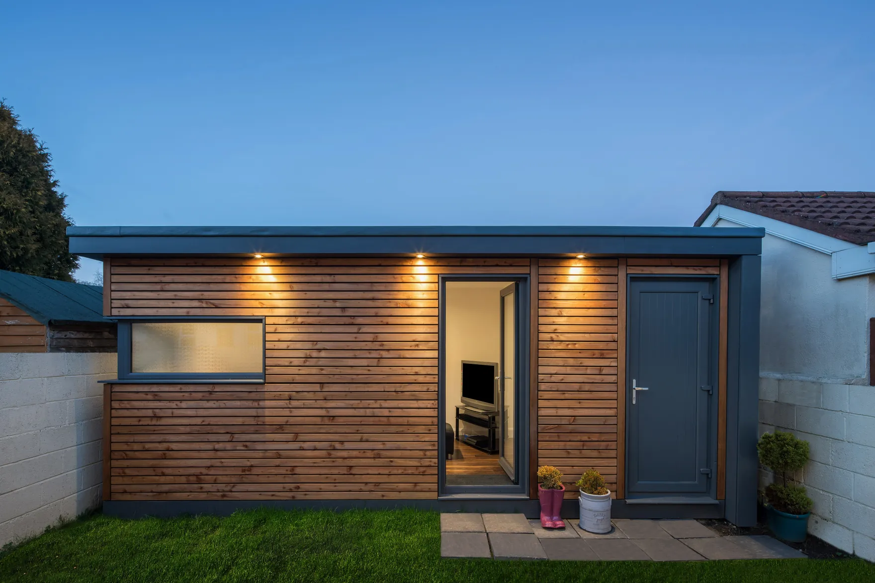 A timber-clad garden room with a grey door, between two boundary walls, lit at dusk