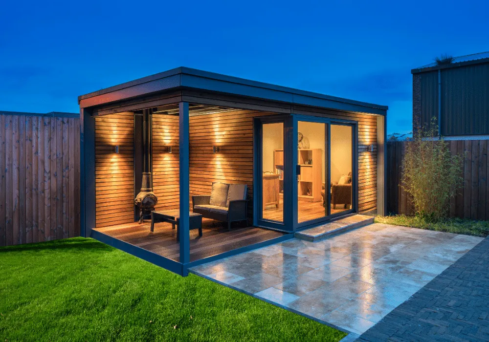 A garden room with a covered timber deck, an outdoor stove and sliding glass doors beside a paved patio, at dusk