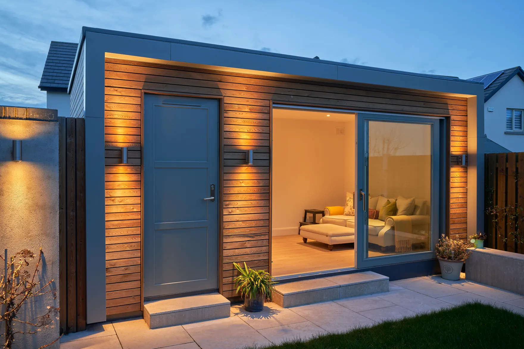 A timber-clad garden room with a grey door and sliding glass doors, lit at dusk