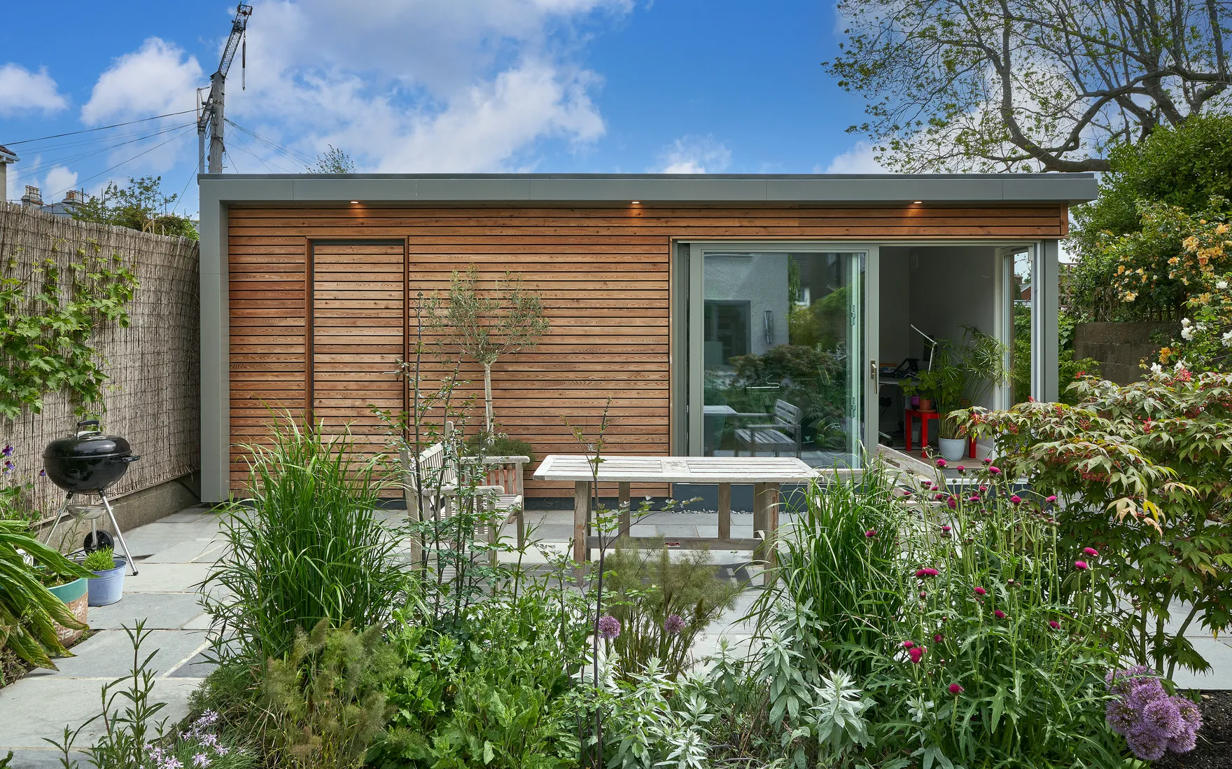 A timber-clad garden room with sliding glass doors behind a planted garden and a patio table