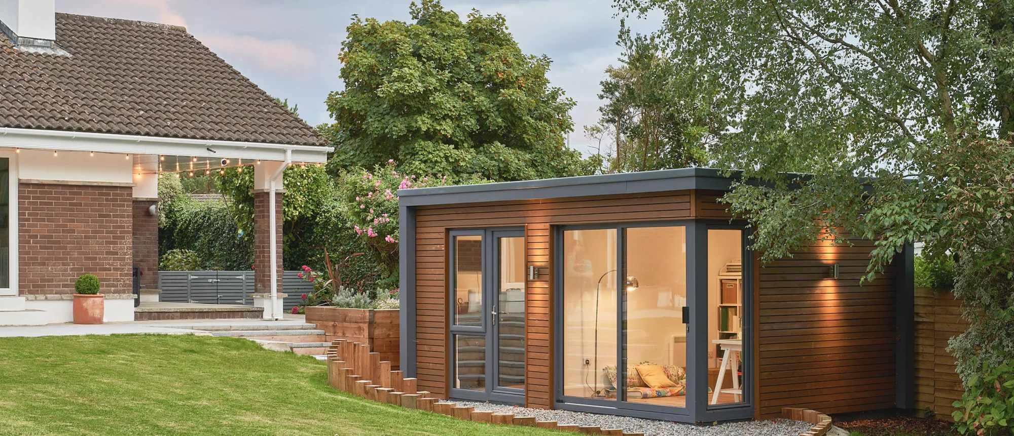 A timber-clad garden room with glass doors in a back garden, beside a house with a covered patio