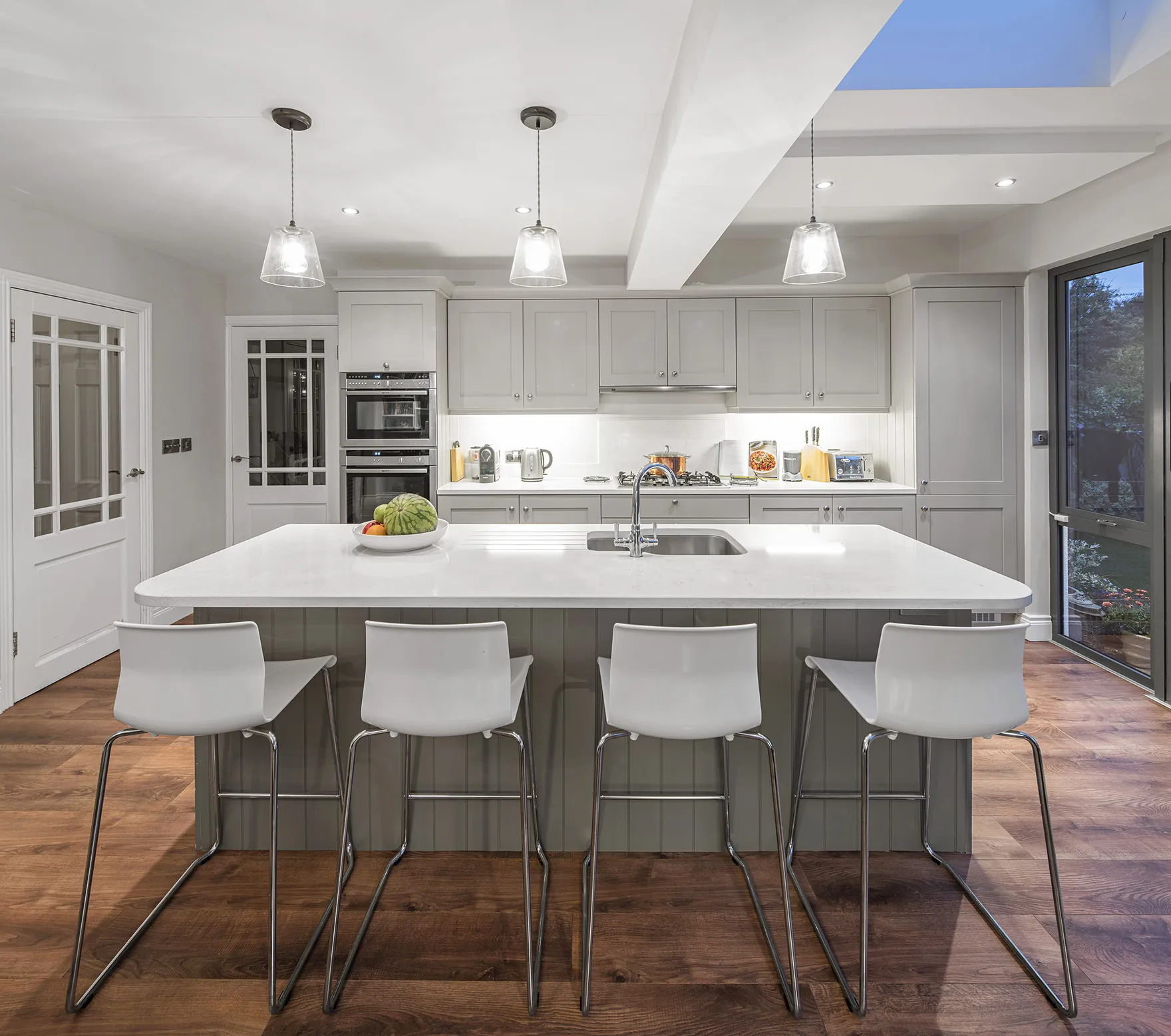A kitchen island with four white bar stools under three glass pendant lights