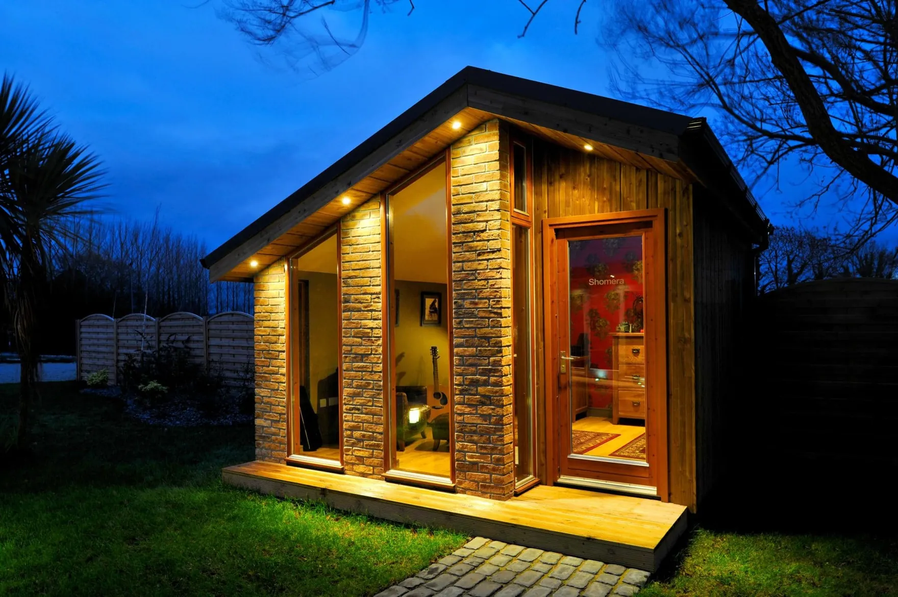 A small garden room with brick piers, tall windows and a pitched roof, lit at dusk
