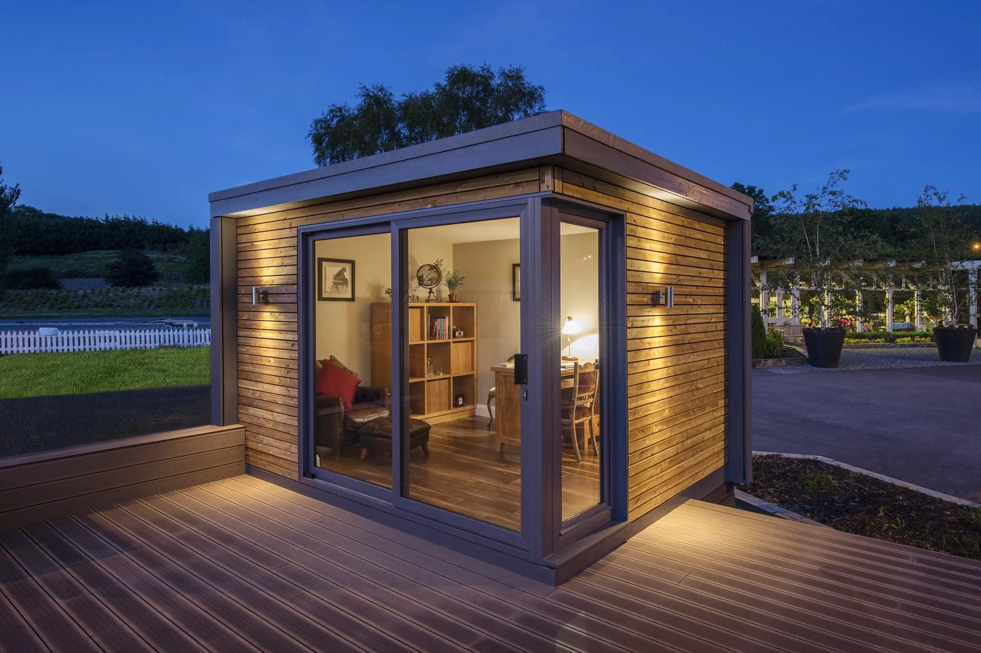 A small timber-clad garden room with sliding glass doors on a deck, lit at dusk