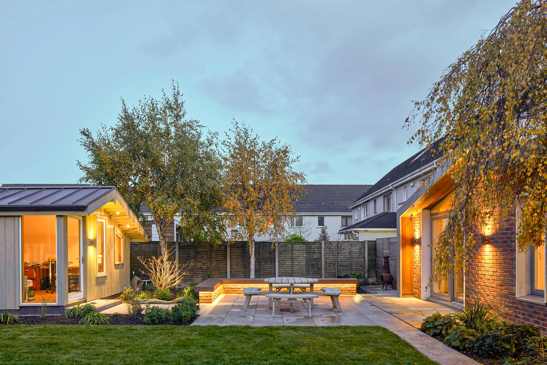 A garden room and a brick house extension around a patio with a picnic table, lit at dusk